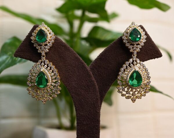 The image shows a pair of elegant drop earrings. The earrings are made with a combination of gold and sparkling gemstones. The main gemstones are teardrop-shaped and appear to be a deep green color, possibly emeralds or moissanites. These large teardrop gems are surrounded by a halo of smaller, white gemstones, likely diamonds or cubic zirconia. The earrings are showcased on a brown jewelry display stand against a backdrop of a potted plant with lush green leaves.
