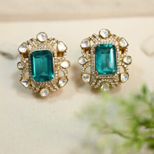 The image shows a pair of elegant earrings with a rectangular emerald-cut gemstone set in a gold frame. The frame is embellished with smaller diamonds or white stones, creating a sparkling halo around the central stone. The earrings are displayed on a white surface with a touch of greenery in the background, suggesting a natural and luxurious setting.