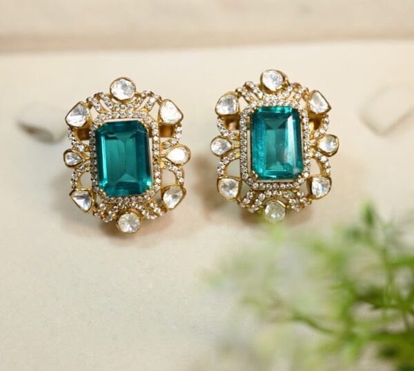 The image shows a pair of elegant earrings with a rectangular emerald-cut gemstone set in a gold frame. The frame is embellished with smaller diamonds or white stones, creating a sparkling halo around the central stone. The earrings are displayed on a white surface with a touch of greenery in the background, suggesting a natural and luxurious setting.