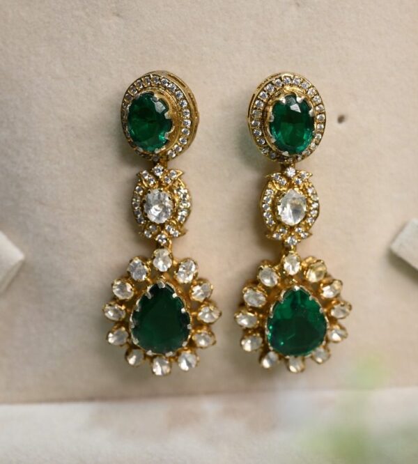 The image shows a pair of elegant drop earrings. They appear to be made of gold and feature a combination of gemstones. There are round, faceted diamonds, possibly white or colorless, and a larger, oval-shaped green stone, which could be an emerald or another green gemstone. The earrings have a delicate, elongated design with multiple drops or pendants, creating a graceful and eye-catching look.