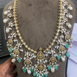The image shows a hand holding a beautiful gold necklace with intricate details. The necklace appears to be made with a combination of gold, pearls, and possibly other gemstones. The central design features a depiction of a deity, likely a Hindu god or goddess. The overall impression is one of elegance, craftsmanship, and cultural significance.