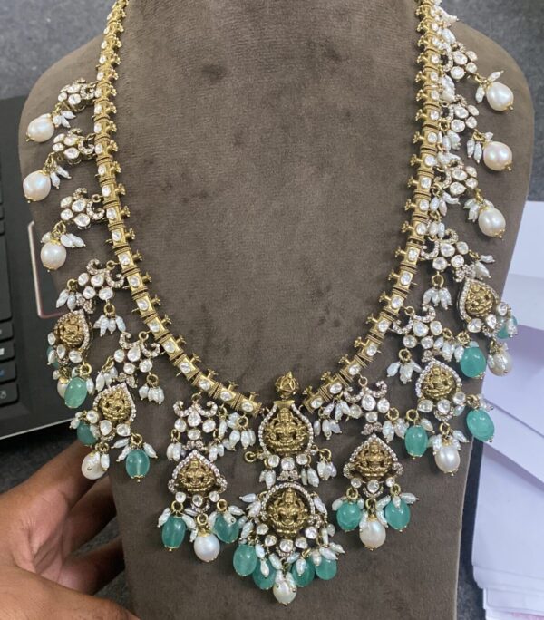 The image shows a hand holding a beautiful gold necklace with intricate details. The necklace appears to be made with a combination of gold, pearls, and possibly other gemstones. The central design features a depiction of a deity, likely a Hindu god or goddess. The overall impression is one of elegance, craftsmanship, and cultural significance.
