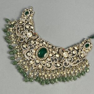 The image shows a close-up of a necklace. The necklace is made of gold and is adorned with numerous gemstones. The central motif of the necklace features a large, round green gemstone surrounded by smaller, white gemstones. The necklace also has several dangling strands of smaller green gemstones. The overall design of the necklace is intricate and ornate.