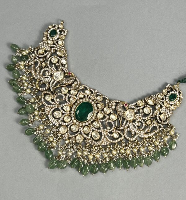 The image shows a close-up of a necklace. The necklace is made of gold and is adorned with numerous gemstones. The central motif of the necklace features a large, round green gemstone surrounded by smaller, white gemstones. The necklace also has several dangling strands of smaller green gemstones. The overall design of the necklace is intricate and ornate.