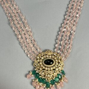 The image shows a multi-strand necklace with pink quartz beads. The necklace features a central pendant adorned with a black onyx stone surrounded by a halo of sparkling moissanites. The pendant is further embellished with vibrant green emeralds and delicate pearls.