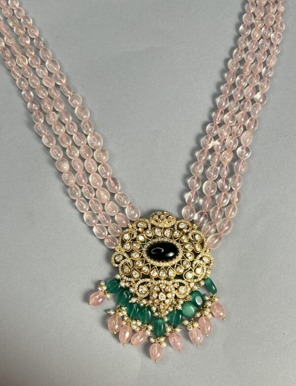 The image shows a multi-strand necklace with pink quartz beads. The necklace features a central pendant adorned with a black onyx stone surrounded by a halo of sparkling moissanites. The pendant is further embellished with vibrant green emeralds and delicate pearls.