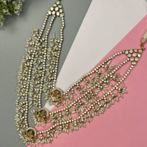 The image shows a close-up of a necklace lying on a surface with contrasting colors (grey and pink). The necklace appears to be made with multiple strands of small, white beads or pearls. There are also larger, round stones interspersed throughout, giving it a luxurious and elegant look. The necklace has a cascading or layered design, with the strands varying in length.