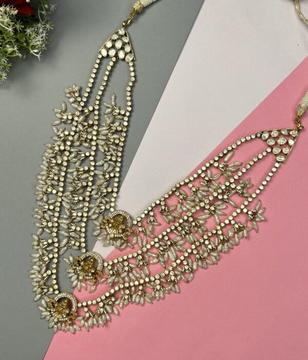 The image shows a close-up of a necklace lying on a surface with contrasting colors (grey and pink). The necklace appears to be made with multiple strands of small, white beads or pearls. There are also larger, round stones interspersed throughout, giving it a luxurious and elegant look. The necklace has a cascading or layered design, with the strands varying in length.