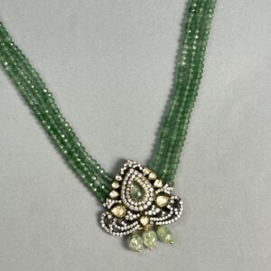 The image shows a close-up of a delicate necklace. It appears to be made with multiple strands of small, faceted green beads. A central pendant hangs from the strands, showcasing a teardrop-shaped design with a cluster of smaller, colorless gemstones set within. The pendant is further embellished with intricate details and possibly additional gemstones. The overall impression is one of elegance and sophistication.