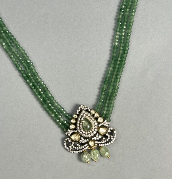 The image shows a close-up of a delicate necklace. It appears to be made with multiple strands of small, faceted green beads. A central pendant hangs from the strands, showcasing a teardrop-shaped design with a cluster of smaller, colorless gemstones set within. The pendant is further embellished with intricate details and possibly additional gemstones. The overall impression is one of elegance and sophistication.