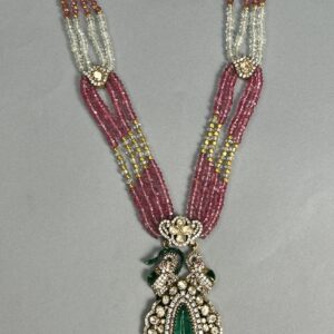 The image shows a beautiful necklace with a large, pear-shaped green moissanite gemstone as the centerpiece. The stone is surrounded by smaller diamonds and pearls, creating a stunning and elegant look. The necklace itself consists of multiple strands of pink and white beads, giving it a layered and luxurious appearance. The overall design is reminiscent of traditional Indian jewelry with a modern twist.