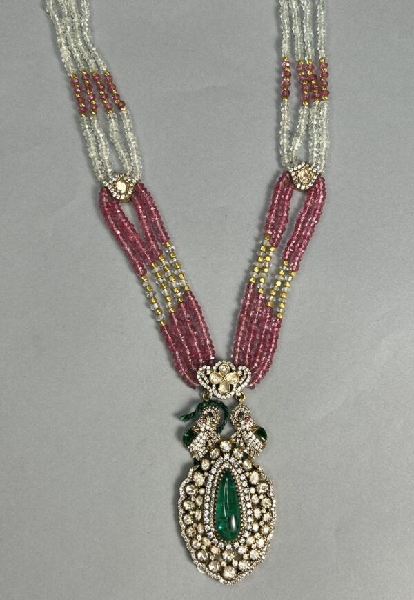 The image shows a beautiful necklace with a large, pear-shaped green moissanite gemstone as the centerpiece. The stone is surrounded by smaller diamonds and pearls, creating a stunning and elegant look. The necklace itself consists of multiple strands of pink and white beads, giving it a layered and luxurious appearance. The overall design is reminiscent of traditional Indian jewelry with a modern twist.