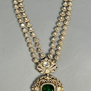 The image shows a necklace. It appears to be made of gold with numerous small, clear stones set into the metal. The necklace has a double strand design, with two parallel chains connected by a series of decorative elements. A larger, oval-shaped pendant hangs from the center of the necklace. This pendant is also adorned with clear stones and features a larger, green stone in the center.