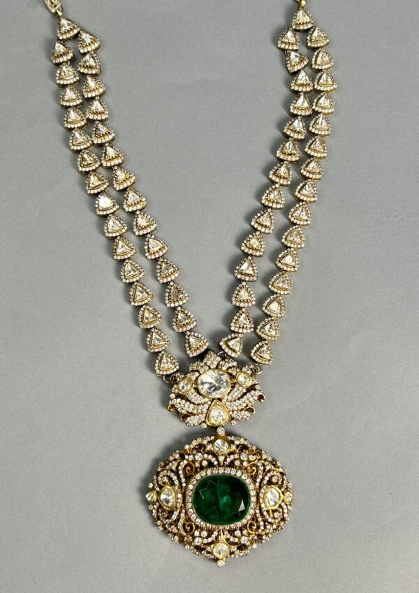 The image shows a necklace. It appears to be made of gold with numerous small, clear stones set into the metal. The necklace has a double strand design, with two parallel chains connected by a series of decorative elements. A larger, oval-shaped pendant hangs from the center of the necklace. This pendant is also adorned with clear stones and features a larger, green stone in the center.