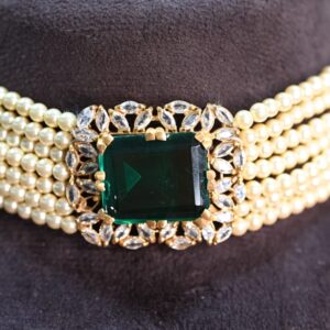 The image shows a close-up of a necklace resting on a dark surface. The necklace has multiple strands of white pearls creating a choker style. At the center, a large square-cut green gemstone is prominently displayed. The gemstone is surrounded by a halo of smaller, sparkling white stones. The overall impression is one of elegance and luxury.