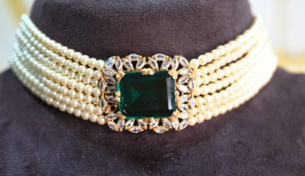 The image shows a close-up of a necklace resting on a dark surface. The necklace has multiple strands of white pearls creating a choker style. At the center, a large square-cut green gemstone is prominently displayed. The gemstone is surrounded by a halo of smaller, sparkling white stones. The overall impression is one of elegance and luxury.