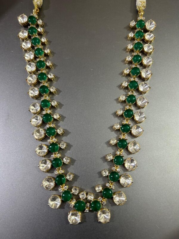 The image shows a necklace. It appears to be made of gold and features a repeating pattern of round, faceted gemstones. The gemstones alternate between a clear color and a deep green. The necklace has a long, flowing design with multiple strands. Unique Moissanite Necklace