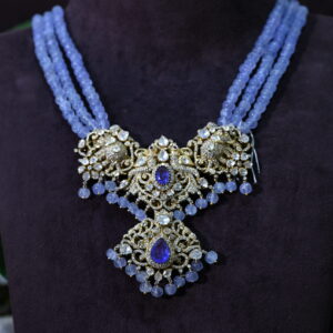 The image shows a close-up of a necklace on a black mannequin. The necklace is made with blue beads and features a large, ornate pendant in the center. The pendant is adorned with intricate gold filigree work and several small, sparkling gemstones. Moissanite