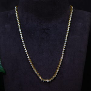 The image shows a necklace with a classic tennis chain design. The chain is adorned with round-cut moissanite stones, creating a continuous line of sparkling brilliance. The necklace appears to be made of gold-plated brass and is displayed on a black mannequin's neck against a dark background. A green plant with large leaves is partially visible in the background.