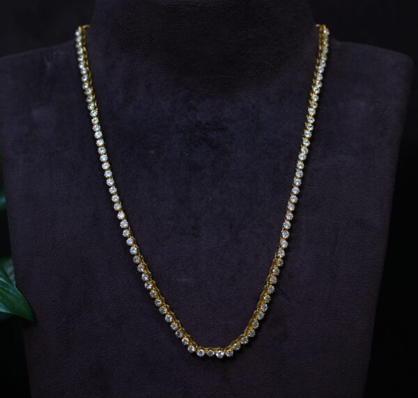 The image shows a necklace with a classic tennis chain design. The chain is adorned with round-cut moissanite stones, creating a continuous line of sparkling brilliance. The necklace appears to be made of gold-plated brass and is displayed on a black mannequin's neck against a dark background. A green plant with large leaves is partially visible in the background.