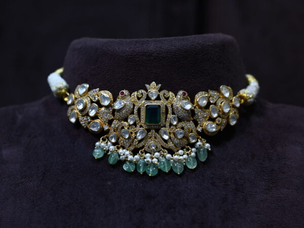 The image shows a necklace on a black velvet stand. The necklace has a central large emerald-cut green gemstone surrounded by smaller white gemstones and pearls. The design appears to be inspired by nature, with intricate floral and leaf motifs. The overall effect is one of luxury and elegance.