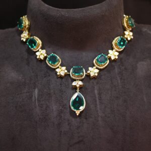 The image shows a close-up of a necklace lying on a dark, velvety surface. The necklace is made of gold and features several large, emerald-green gemstones. The central gemstone is a pear-shaped pendant, and it is surrounded by smaller, square-cut gemstones. The necklace also incorporates delicate gold filigree work and smaller, flower-shaped motifs with pavé-set gemstones. The overall effect is luxurious and elegant. Premium Moissanite Necklace