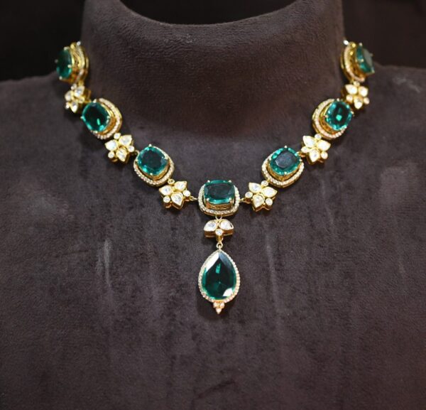 The image shows a close-up of a necklace lying on a dark, velvety surface. The necklace is made of gold and features several large, emerald-green gemstones. The central gemstone is a pear-shaped pendant, and it is surrounded by smaller, square-cut gemstones. The necklace also incorporates delicate gold filigree work and smaller, flower-shaped motifs with pavé-set gemstones. The overall effect is luxurious and elegant. Premium Moissanite Necklace