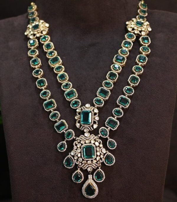 The image shows a beautiful necklace with a double-layered design. It's adorned with numerous emerald-cut moissanites, which are known for their exceptional brilliance and fire. The stones are set in a delicate gold frame, creating a luxurious and sophisticated look. The necklace features a central pendant with a larger moissanite surrounded by smaller stones. The pendant is connected to the double-layered chain, which is also adorned with emerald-cut moissanites throughout.
