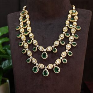 The necklace has multiple strands with alternating emerald-cut moissanites and round brilliant-cut diamonds. The central motif features a larger emerald-cut moissanite surrounded by a halo of smaller diamonds. The necklace appears to be resting on a dark surface with a small potted plant in the background.