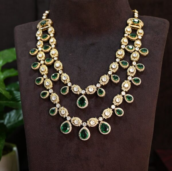 The necklace has multiple strands with alternating emerald-cut moissanites and round brilliant-cut diamonds. The central motif features a larger emerald-cut moissanite surrounded by a halo of smaller diamonds. The necklace appears to be resting on a dark surface with a small potted plant in the background.
