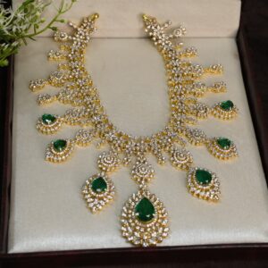 The image shows a beautiful necklace with a cascading design. It appears to be made of gold and is adorned with numerous small, sparkling stones, likely diamonds or cubic zirconia. Several larger, pear-shaped emerald green gemstones are also visible, adding a pop of color. The necklace is resting on a luxurious-looking velvet or satin surface, possibly inside a jewelry box.