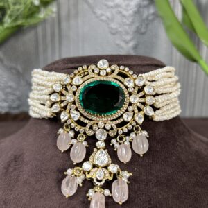 The image shows a beautiful necklace. It has a large, oval-shaped green gemstone in the center, likely an emerald. The emerald is surrounded by smaller, sparkling white gemstones, possibly diamonds or moissanites. These smaller stones are set in intricate patterns, creating a dazzling effect. The necklace also features a delicate chain of pearls and has a dangling pendant with pink gemstones. The overall style is elegant and luxurious.