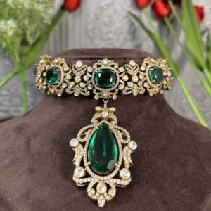 The image shows a close-up of a necklace lying on a dark, velvety surface. The necklace has a wide band encrusted with numerous small, round, colorless stones. From the center of this band, a large, pear-shaped, deep green stone dangles on a delicate, ornate chain. The chain is also decorated with small, round, colorless stones. The background is a blurred image of red tulips with green stems.