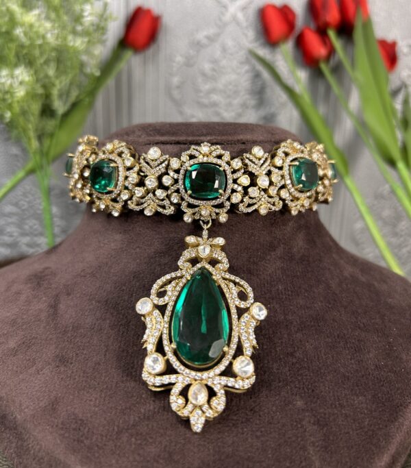The image shows a close-up of a necklace lying on a dark, velvety surface. The necklace has a wide band encrusted with numerous small, round, colorless stones. From the center of this band, a large, pear-shaped, deep green stone dangles on a delicate, ornate chain. The chain is also decorated with small, round, colorless stones. The background is a blurred image of red tulips with green stems.