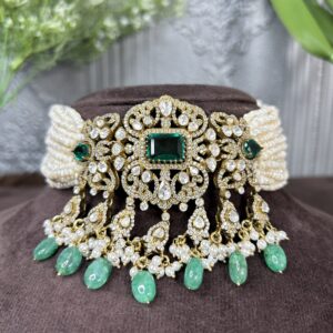 The image shows a close-up of a necklace lying on a brown surface. The necklace appears to be made of gold or a gold-toned metal and is adorned with a large, ornate centerpiece. This centerpiece features a square emerald gemstone surrounded by a intricate design of smaller diamonds or moissanite stones.