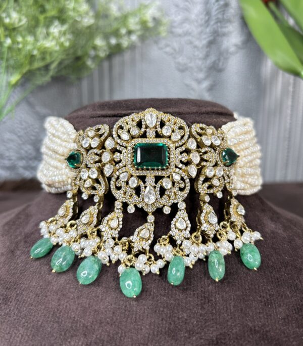 The image shows a close-up of a necklace lying on a brown surface. The necklace appears to be made of gold or a gold-toned metal and is adorned with a large, ornate centerpiece. This centerpiece features a square emerald gemstone surrounded by a intricate design of smaller diamonds or moissanite stones.
