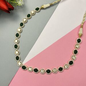 The image shows a necklace with a delicate chain and a row of oval-shaped green stones. The stones are surrounded by smaller, sparkling white stones, possibly diamonds or cubic zirconia. The necklace has a gold-tone finish and appears to be made of metal. The background is a combination of gray, white, and pink surfaces, with a red flower visible in the top left corner.
