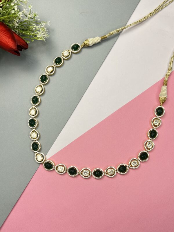 The image shows a necklace with a delicate chain and a row of oval-shaped green stones. The stones are surrounded by smaller, sparkling white stones, possibly diamonds or cubic zirconia. The necklace has a gold-tone finish and appears to be made of metal. The background is a combination of gray, white, and pink surfaces, with a red flower visible in the top left corner.