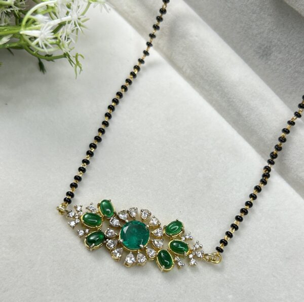 A gold necklace with black beads and a green stone pendant surrounded by smaller white stones. Close-up of a necklace with a central emerald stone and diamond accents.