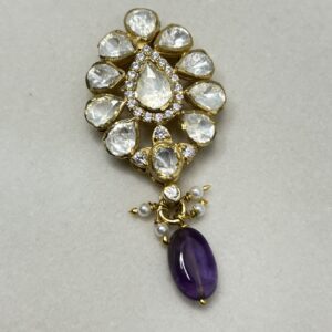 The image shows a close-up of a delicate pendant. It appears to be made of gold and is adorned with a cluster of sparkling white stones, likely diamonds or moissanites. The stones are arranged in a flower-like pattern, with a larger stone in the center. A single purple gemstone, possibly amethyst, hangs from the bottom of the pendant, adding a touch of color. The pendant is resting on a white surface, possibly a piece of cloth or a jewelry display.