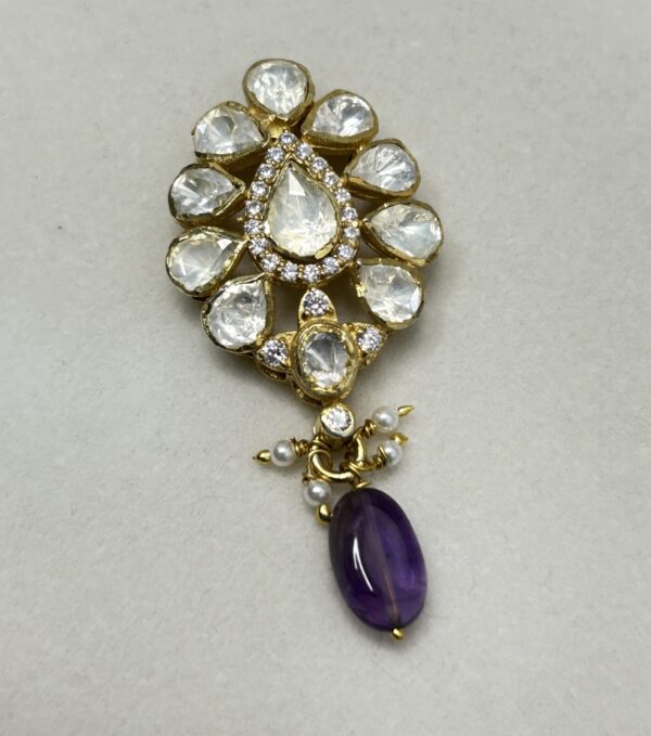 The image shows a close-up of a delicate pendant. It appears to be made of gold and is adorned with a cluster of sparkling white stones, likely diamonds or moissanites. The stones are arranged in a flower-like pattern, with a larger stone in the center. A single purple gemstone, possibly amethyst, hangs from the bottom of the pendant, adding a touch of color. The pendant is resting on a white surface, possibly a piece of cloth or a jewelry display.