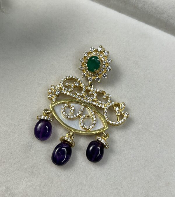 "A silver pendant with a filigree eye design, accented with green and purple gemstones." "A close-up of an ornate silver pendant with moissanites, a green stone, and amethyst drops."