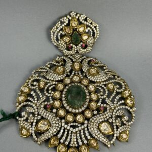 The image shows a close-up of a large, ornate pendant. It appears to be made of gold or a similar metal and is heavily embellished with gemstones. A large, central green gemstone is surrounded by numerous smaller, colorless gemstones, likely diamonds or moissanites. The overall design is intricate and symmetrical, with delicate filigree work creating a floral motif. The pendant seems to be suspended from a thin chain.