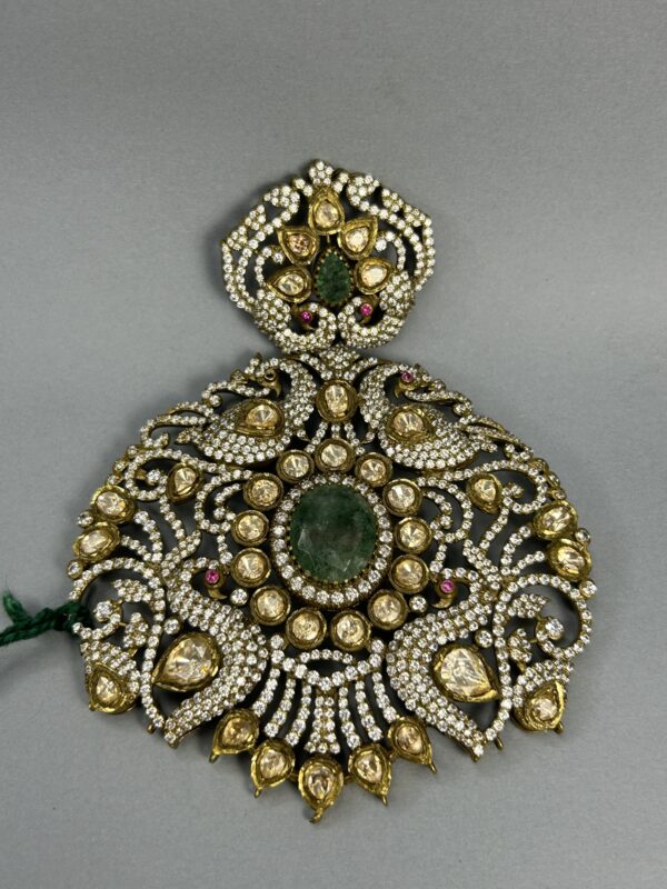 The image shows a close-up of a large, ornate pendant. It appears to be made of gold or a similar metal and is heavily embellished with gemstones. A large, central green gemstone is surrounded by numerous smaller, colorless gemstones, likely diamonds or moissanites. The overall design is intricate and symmetrical, with delicate filigree work creating a floral motif. The pendant seems to be suspended from a thin chain.