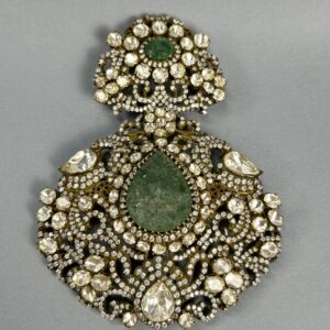 The image shows a close-up of a large, ornate pendant. It appears to be made of gold or a similar metal and is heavily embellished with gemstones. A large, central green gemstone is surrounded by numerous smaller, colorless gemstones, likely diamonds or moissanites. The overall design is intricate and symmetrical, with delicate filigree work creating a floral motif. The pendant seems to be suspended from a thin chain.