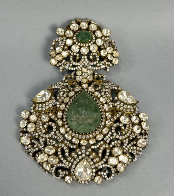 The image shows a close-up of a large, ornate pendant. It appears to be made of gold or a similar metal and is heavily embellished with gemstones. A large, central green gemstone is surrounded by numerous smaller, colorless gemstones, likely diamonds or moissanites. The overall design is intricate and symmetrical, with delicate filigree work creating a floral motif. The pendant seems to be suspended from a thin chain.