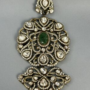 The image shows a close-up of a large, ornate pendant. It appears to be made of gold or a similar metal and is heavily embellished with gemstones. A large, central green gemstone is surrounded by numerous smaller, colorless gemstones, likely diamonds or moissanites. The overall design is intricate and symmetrical, with delicate filigree work creating a floral motif. The pendant seems to be suspended from a thin chain.
