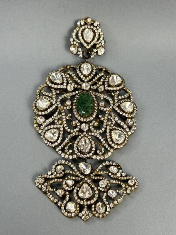 The image shows a close-up of a large, ornate pendant. It appears to be made of gold or a similar metal and is heavily embellished with gemstones. A large, central green gemstone is surrounded by numerous smaller, colorless gemstones, likely diamonds or moissanites. The overall design is intricate and symmetrical, with delicate filigree work creating a floral motif. The pendant seems to be suspended from a thin chain.