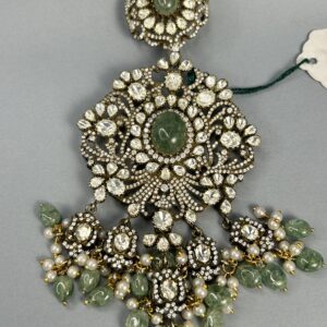 Color: The central stone is a vibrant green, while the surrounding stones are colorless, likely diamonds or moissanites. The metal appears to be silver or white gold. Style: The jewelry piece has a luxurious and elegant look, with its intricate design and use of high-quality gemstones. Size: The pendant appears to be of medium size, suitable for everyday wear.