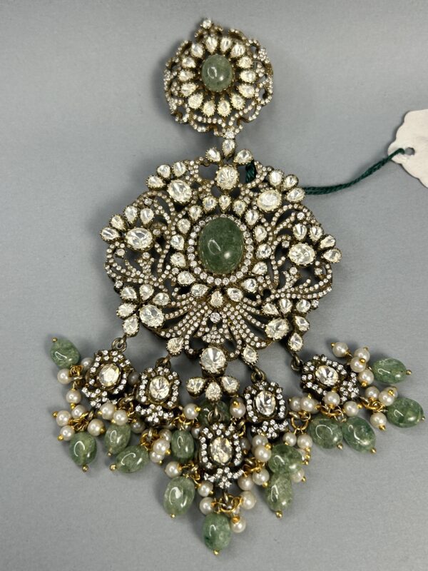 Color: The central stone is a vibrant green, while the surrounding stones are colorless, likely diamonds or moissanites. The metal appears to be silver or white gold. Style: The jewelry piece has a luxurious and elegant look, with its intricate design and use of high-quality gemstones. Size: The pendant appears to be of medium size, suitable for everyday wear.